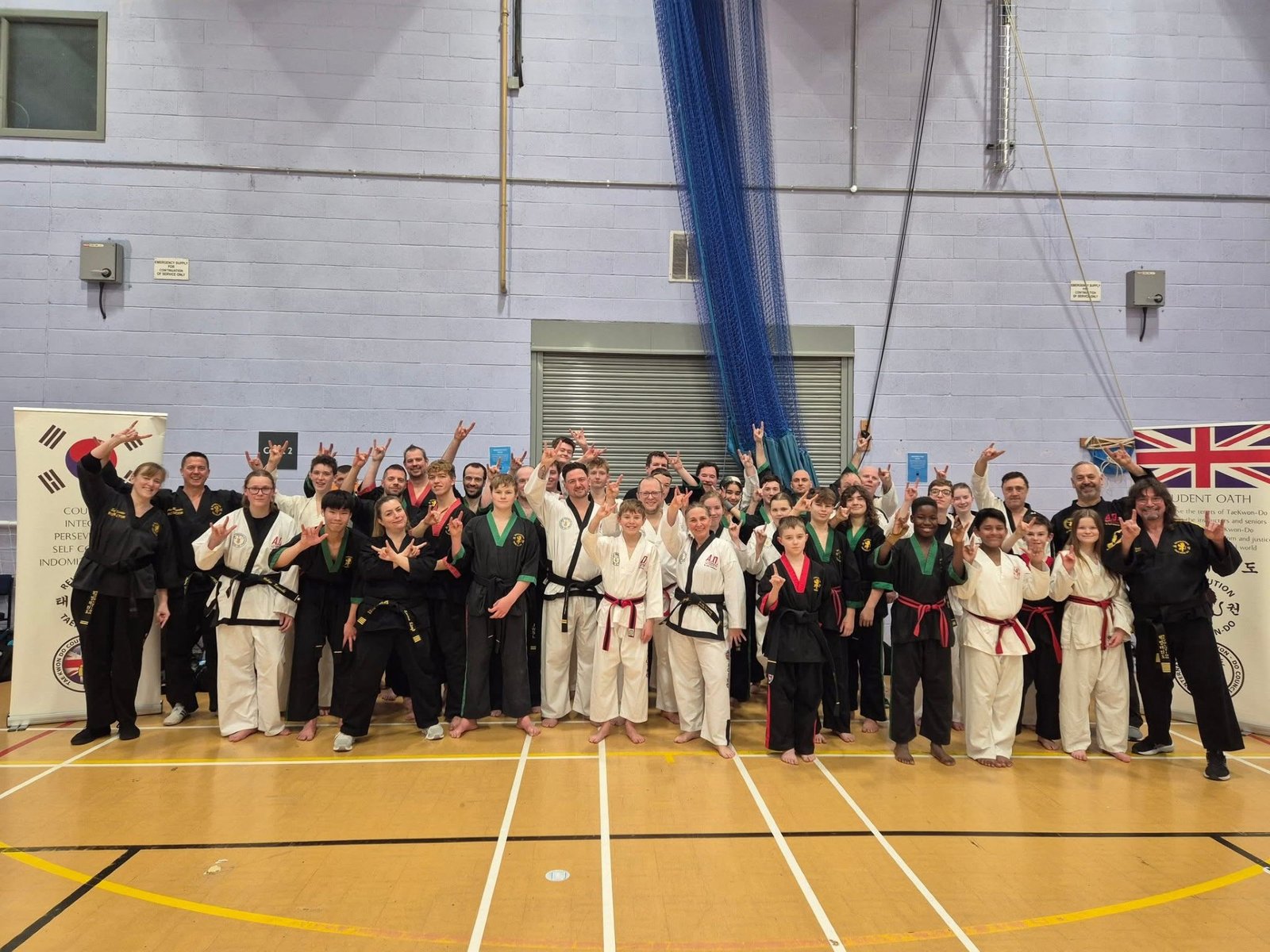 A large group of martial artists in black and white uniforms posing together in a sports hall, smiling and holding up hand signs.